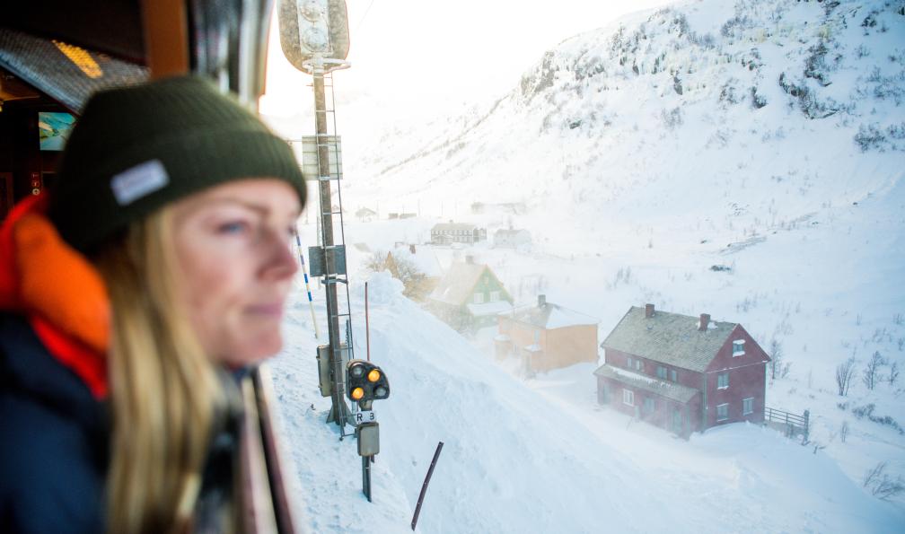Hjørnevik - Sverre - Norway's Best AS - Femme dans le train en hiver qui contemple les montagnes enneigées et les maisons traditionnelles rouges à Flamsbana, Norvège - Découverte hivernale des fjords norvégiens - 9